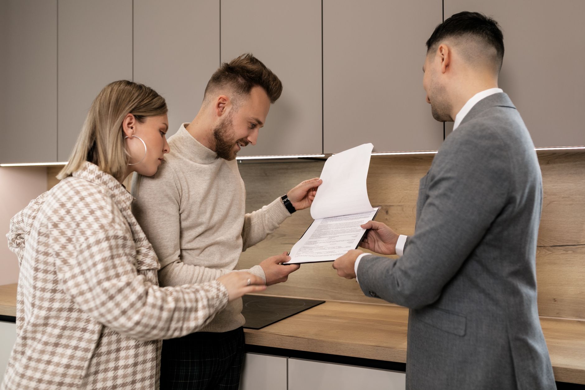 A couple and an agent in a modern kitchen looking over a printed document together.