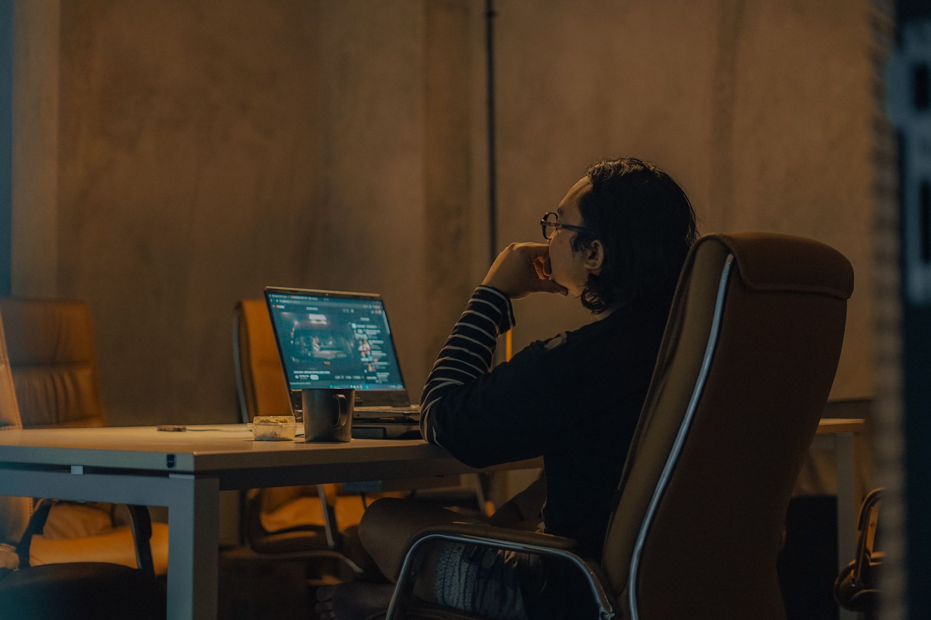 A man sits at a desk working on his laptop late in a dimly lit home office, with a cup of coffee nearby.
